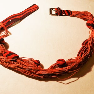 May include: A handcrafted necklace with orange threads and fabric details. The fabric is red with black stripes. The necklace has a buckle fastening and several round fabric beads.