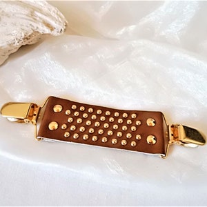 May include: A brown leather garment clip with gold metal accents and a studded design.