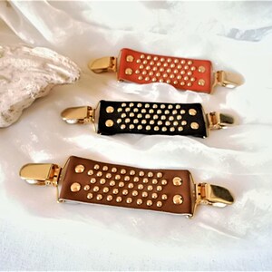 May include: Three leather hair clips with gold metal accents. The clips are brown, black, and orange and have a studded design.