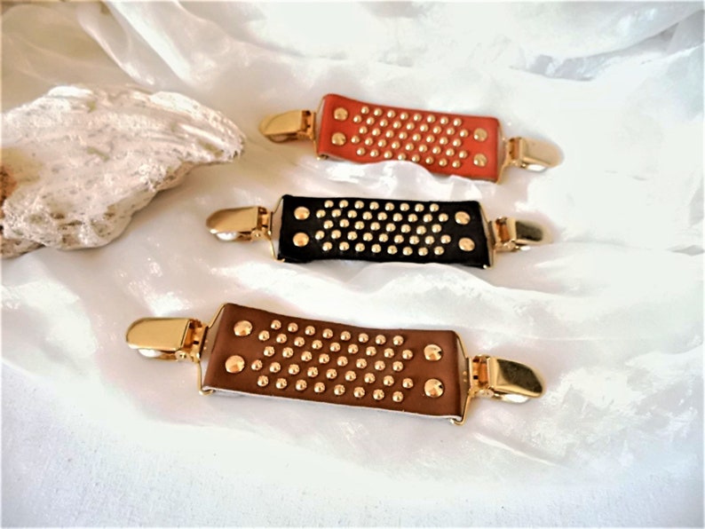 May include: Three leather hair clips with gold metal accents. The clips are brown, black, and orange and have a studded design.
