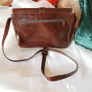 May include: A brown leather crossbody bag with a flap closure and a zippered pocket on the back. The bag has a long adjustable strap.