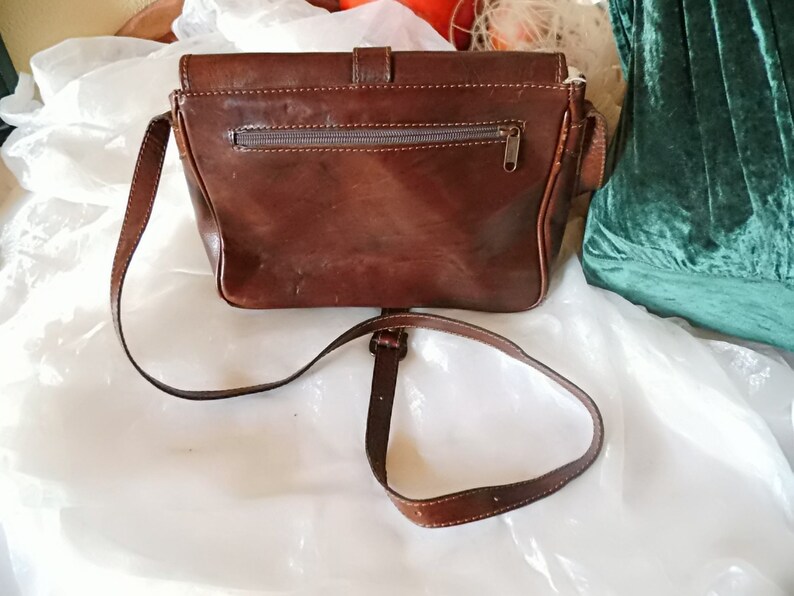 May include: A brown leather crossbody bag with a flap closure and a zippered pocket on the back. The bag has a long adjustable strap.