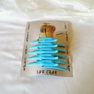 Könnte beinhalten: Eine Packung blauer Plastik-Haarklammern namens "The Crab". Die Verpackung zeigt ein Model mit blonden Haaren, das die Klammern trägt.