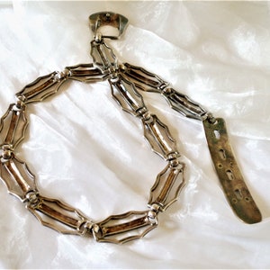 May include: A silver chain belt with a decorative buckle and a long, flat, silver end piece. The chain is made of interlocking links with a geometric design.