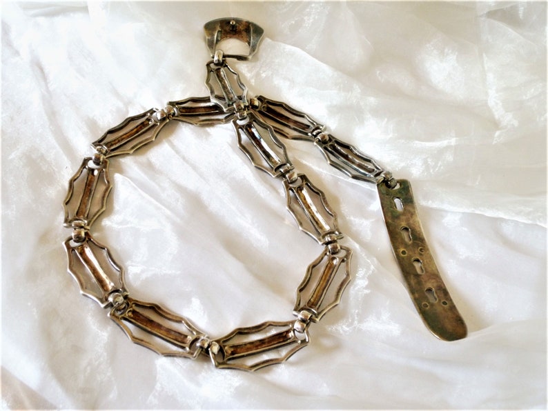 May include: A silver chain belt with a decorative buckle and a long, flat, silver end piece. The chain is made of interlocking links with a geometric design.