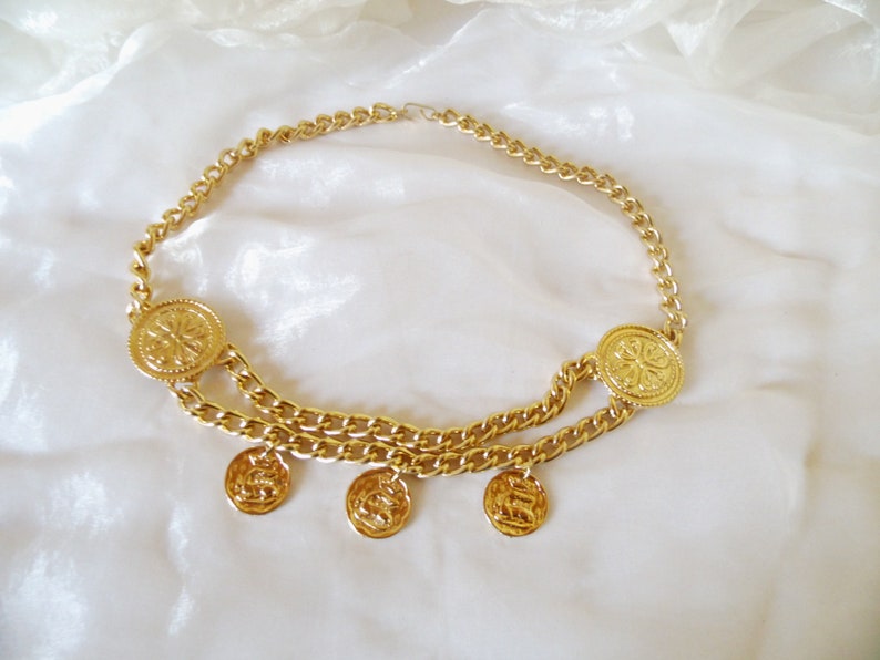 May include: A gold chain belt with large round coin-shaped charms. The belt has two chains and four dangling charms.