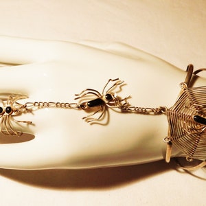 Könnte beinhalten: Ein goldenes Armband mit einem Spinnennetz-Design und zwei baumelnden Spinnenanhängern. Das Armband ist aus Draht gefertigt und hat schwarze Onyxsteine für die Augen der Spinnen.