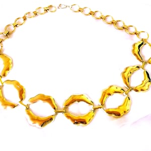 Könnte beinhalten: Eine goldfarbene Metallkettenhalskette mit einer Reihe von miteinander verbundenen kreisförmigen Gliedern. Der obere Teil besteht aus einfachen runden Ringen, während der untere Abschnitt größere, organische, wellenförmige Kreise aufweist.