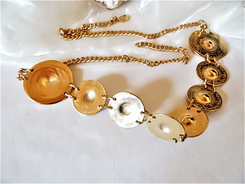 Puede incluir: Cintur&oacute;n de cadena dorada con m&uacute;ltiples dijes redondos, decorativos y con forma de moneda.