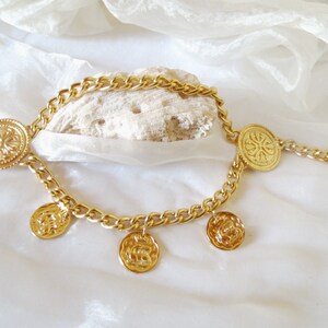 May include: Gold chain necklace with multiple round coin-shaped pendants. The chain is thick and has a curb link design. The pendants have a raised floral design.