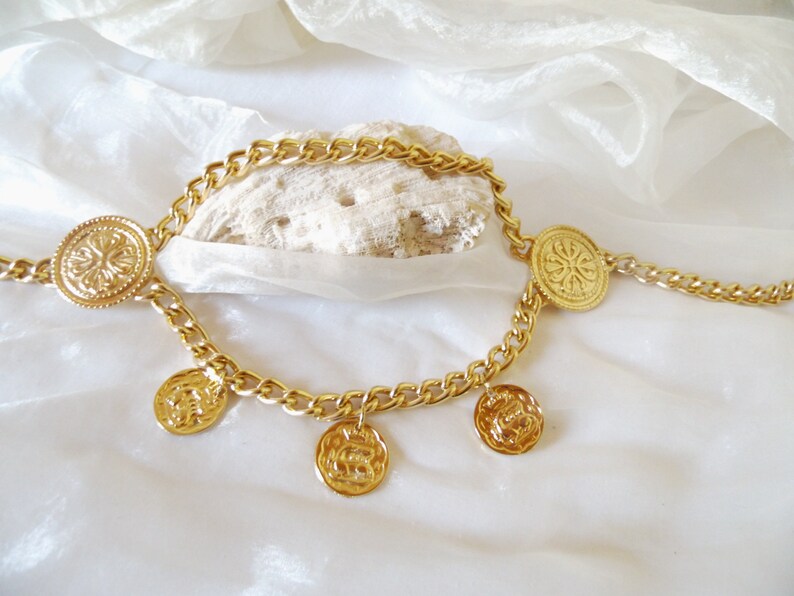 May include: Gold chain necklace with multiple round coin-shaped pendants. The chain is thick and has a curb link design. The pendants have a raised floral design.