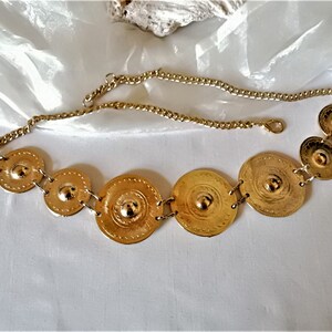 Puede incluir: Un cintur&oacute;n de cadena dorado con seis placas redondas de metal decorativas. Las placas est&aacute;n conectadas por la cadena y tienen un centro elevado.