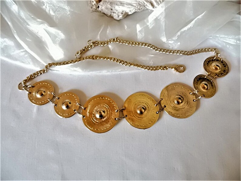 Puede incluir: Un cintur&oacute;n de cadena dorado con seis placas redondas de metal decorativas. Las placas est&aacute;n conectadas por la cadena y tienen un centro elevado.