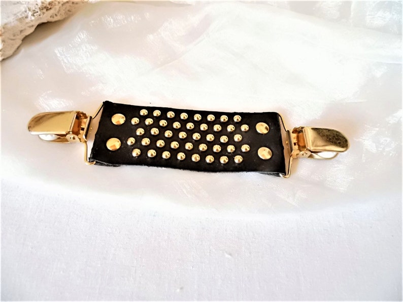 May include: A black leather suspender clip with gold-toned metal clips and studs.