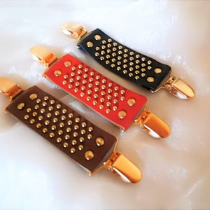 May include: Three leather garment clips with gold-toned metal clasps and studded with gold-toned metal dots. The clips are in black, red, and brown.