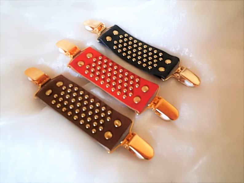 May include: Three leather garment clips with gold-toned metal clasps and studded with gold-toned metal dots. The clips are in black, red, and brown.