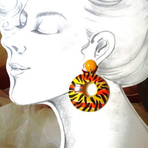 Op de afbeelding: Een paar ronde houten oorbellen in oranje, geel en zwart met een gat in het midden. De oorbellen hangen aan een kleine oranje kraal.