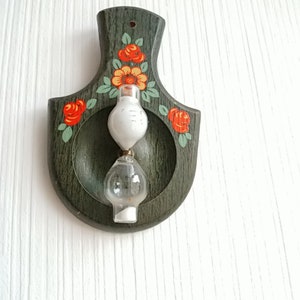 Può includere: Una clessidra di legno con uno sfondo dipinto di verde e motivi floreali rossi e arancioni. La clessidra è riempita di sabbia bianca ed è montata su un muro.