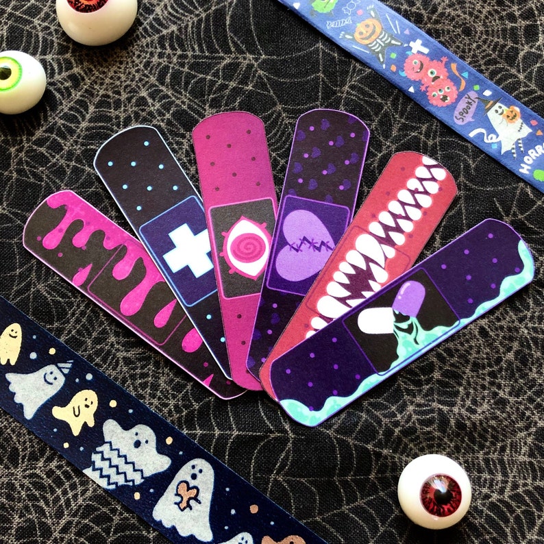 Peut inclure: Une collection de pansements d&eacute;coratifs sur le th&egrave;me d'Halloween. Les pansements pr&eacute;sentent des illustrations de sang qui coule, d'une croix, d'un c&oelig;ur, de dents et d'une pilule. Deux rubans d&eacute;coratifs avec des motifs de fant&ocirc;mes et de monstres sont &eacute;galement visibles.