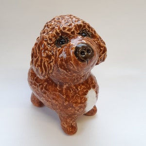 May include: A brown ceramic dog figurine with curly hair and a white patch on its chest. The dog has a realistic face with brown eyes and a black nose.