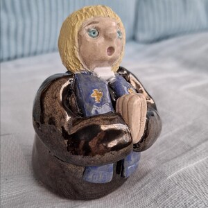 May include: A ceramic figurine depicting a person with blonde hair, blue eyes, and an open mouth. The figure is dressed in a dark brown robe, a blue garment with a gold cross, and holds a light-coloured object. The figurine has a surprised expression.