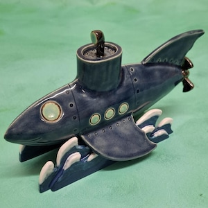 May include: A ceramic submarine sculpture in shades of blue with white accents. The whimsical design features a periscope, portholes, and fins. The submarine has a propeller and is designed to sit on a flat surface.