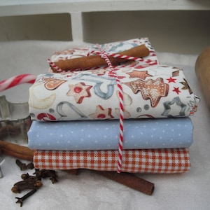 May include: A stack of four fabric squares, tied together with a red and white twine. The top fabric square has a white background with a pattern of gingerbread cookies, candy canes, and stars. The second fabric square is light blue with white polka dots. The third fabric square is orange and white gingham. The bottom fabric square is a solid orange color.