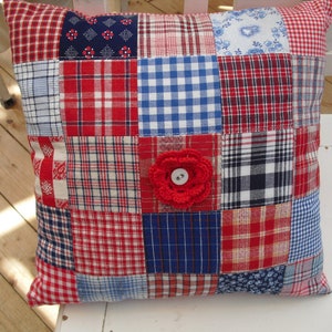 Puede incluir: Una almohada de patchwork rojo, blanco y azul con una flor roja de ganchillo en el centro. La almohada está hecha de diferentes telas a cuadros y es perfecta para añadir un toque de encanto rústico a cualquier habitación.