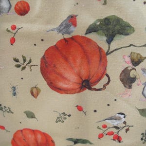Könnte beinhalten: Ein skurriles Stoffdesign mit einer großen orangefarbenen Kürbis, Vögeln und kleinen Figuren. Die Illustration enthält Blätter, Beeren und Insekten auf beigem Hintergrund. Das Gesamtthema ist Herbst und Natur.