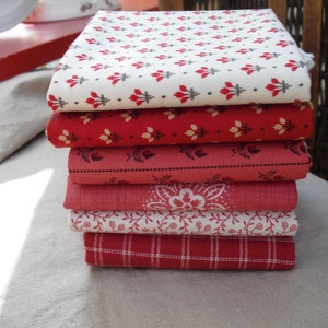 Può includere: Una pila di sei quadrati di tessuto piegati in tonalità di rosso e bianco. I tessuti presentano vari motivi, tra cui floreali, a quadri e geometrici. La pila è appoggiata su una superficie di colore neutro.