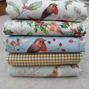 Puede incluir: Conjunto de seis cuadrados de tela con un tema navideño de invierno. Los cuadrados de tela presentan un patrón repetitivo de petirrojos, acebo, bayas y copos de nieve sobre un fondo blanco, azul y marrón.