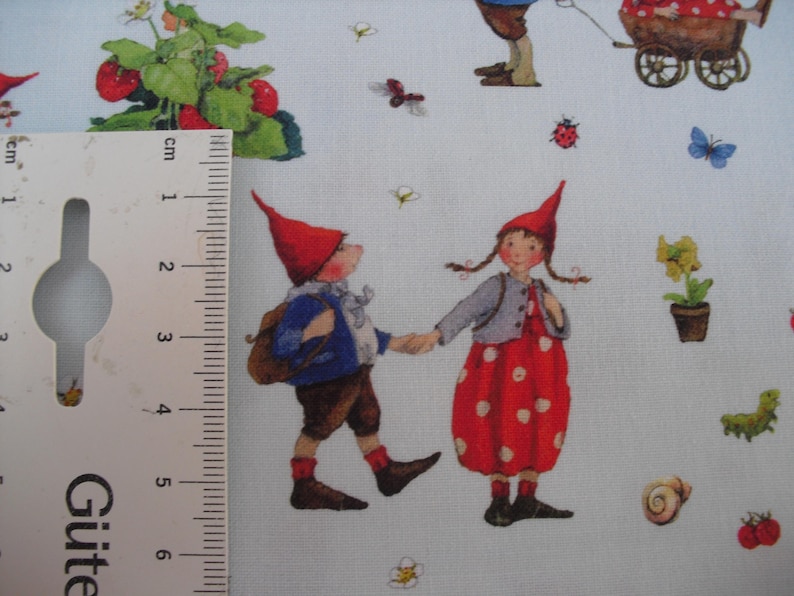 May include: Light blue fabric with a repeating pattern of whimsical gnomes, strawberries, flowers, and insects. The gnomes are depicted in red hats and colorful clothing, holding hands. The fabric is suitable for quilting and crafting projects.