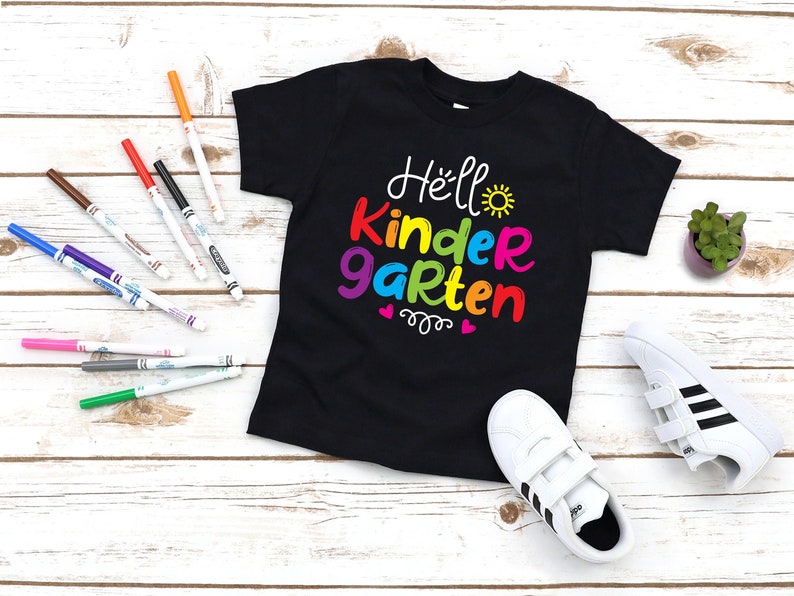 May include: Black t-shirt with the text "Hello Kindergarten" in rainbow colors. The text is in a playful font with a sun and hearts.