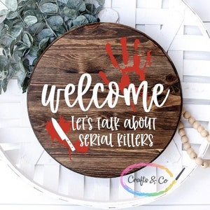 May include: A round wooden sign with a dark brown stain. The sign says "Welcome" in white cursive lettering. Below the welcome greeting, the sign says "Let's talk about serial killers" in white lettering. A red handprint is visible in the upper right corner of the sign. A red knife is visible in the lower left corner of the sign.