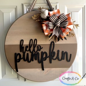 May include: A round wooden door hanger with a brown and white striped bow, a white cotton ball, and a brown and orange plaid bow. The words "hello pumpkin" are written in black on the door hanger.