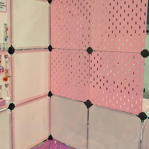 Może przedstawiać: Modułowe stoisko wystawiennicze z różowymi panelami z otworami i białymi panelami. Stoisko jest zbudowane z czarnych łączników i stoi na odblaskowej, opalizującej powierzchni. Płyta perforowana ma małe, równomiernie rozmieszczone otwory.