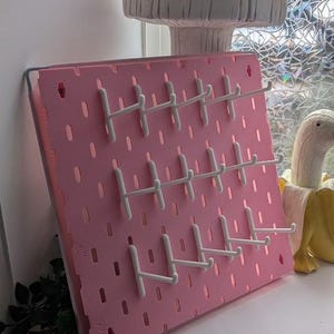May include: A pink pegboard with white hooks. The board is rectangular with evenly spaced holes. A large mushroom decoration is in the background. The pegboard is leaning against a window.