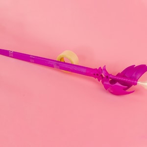 May include: A bright magenta fantasy sword with a detailed hilt and blade, taped to a pink surface. The hilt features a stylized design with wing-like elements. The blade is long and pointed. The sword is approximately 76 cm long.