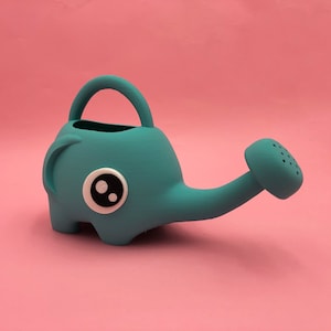 May include: A teal-colored watering can shaped like an elephant with a large, round eye on its side. The can has a curved handle and a spout with small holes for watering plants. The background is a solid pink.