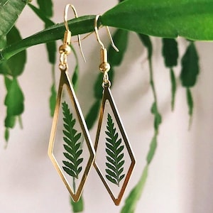 May include: A pair of gold earrings with a diamond-shaped design. Each earring features a pressed fern leaf encased in clear resin.