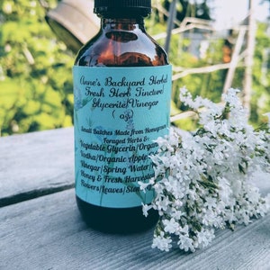 May include: A brown glass bottle with a black dropper top, labeled "Anne's Backyard Herbal Fresh Herb Tincture Glycerite Vinegar (0z)". The bottle is sitting on a wooden surface with white flowers in the background.