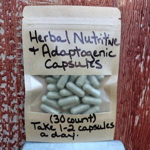Pu&ograve; includere: Una busta di carta marrone con un'etichetta bianca che recita "Herbal Nutritive + Adaptogenic Capsules (30 count) Take 1-2 capsules a day.". La busta contiene un sacchetto di plastica trasparente pieno di capsule verde chiaro.