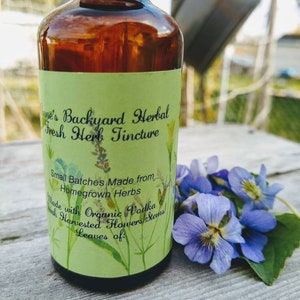 May include: A brown glass bottle with a black cap and a green label featuring text that reads "Anne's Backyard Herbal Fresh Herb Tincture." The label also states "Small Batches Made from Homegrown Herbs." A small bouquet of purple flowers rests beside the bottle.