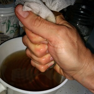 May include: A hand squeezing a white cheesecloth bag over a white ceramic mug filled with a dark brown liquid.