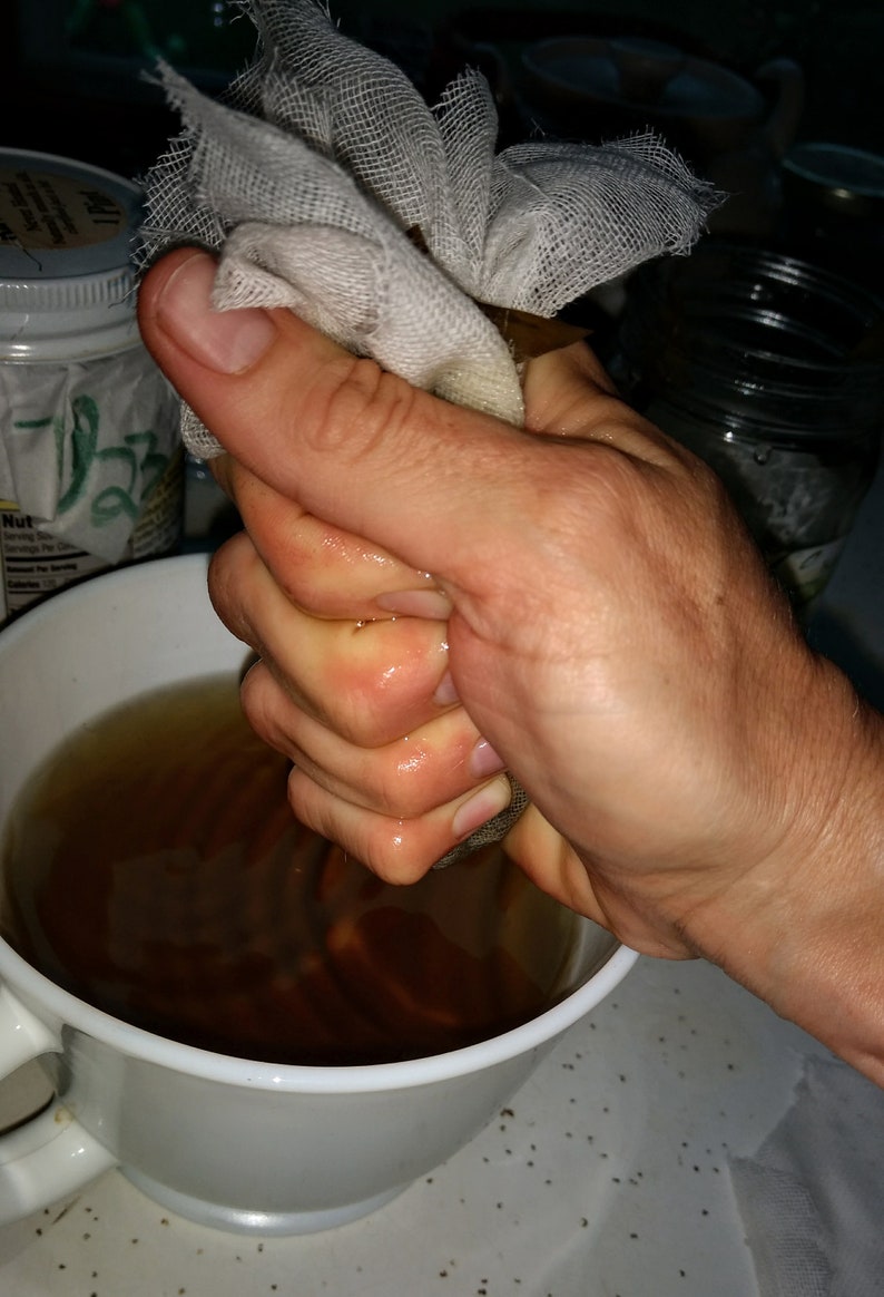 May include: A hand squeezing a white cheesecloth bag over a white ceramic mug filled with a dark brown liquid.