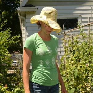 May include: A woman wearing a green t-shirt with a white graphic of potted plants, a straw hat, and blue jeans. She is standing in a garden with green plants.
