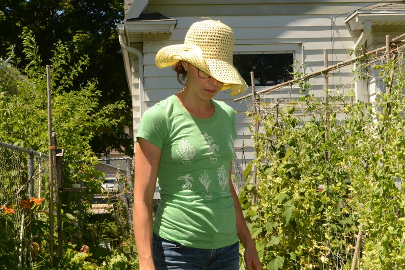 May include: A woman wearing a green t-shirt with a white graphic of potted plants, a straw hat, and blue jeans. She is standing in a garden with green plants.
