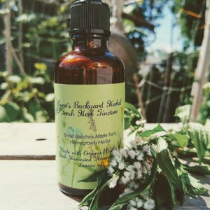 May include: A brown glass bottle with a black dropper, labeled "Anne's Backyard Herbal Fresh Herb Tincture." The label is light green with text and floral illustrations. Fresh herbs and flowers are arranged next to the bottle on a wooden surface.