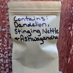 Pu&ograve; includere: Un sacchetto di carta marrone con un'etichetta scritta a mano che dice "Contiene: Tarassaco, Ortica + Ashwagandha."