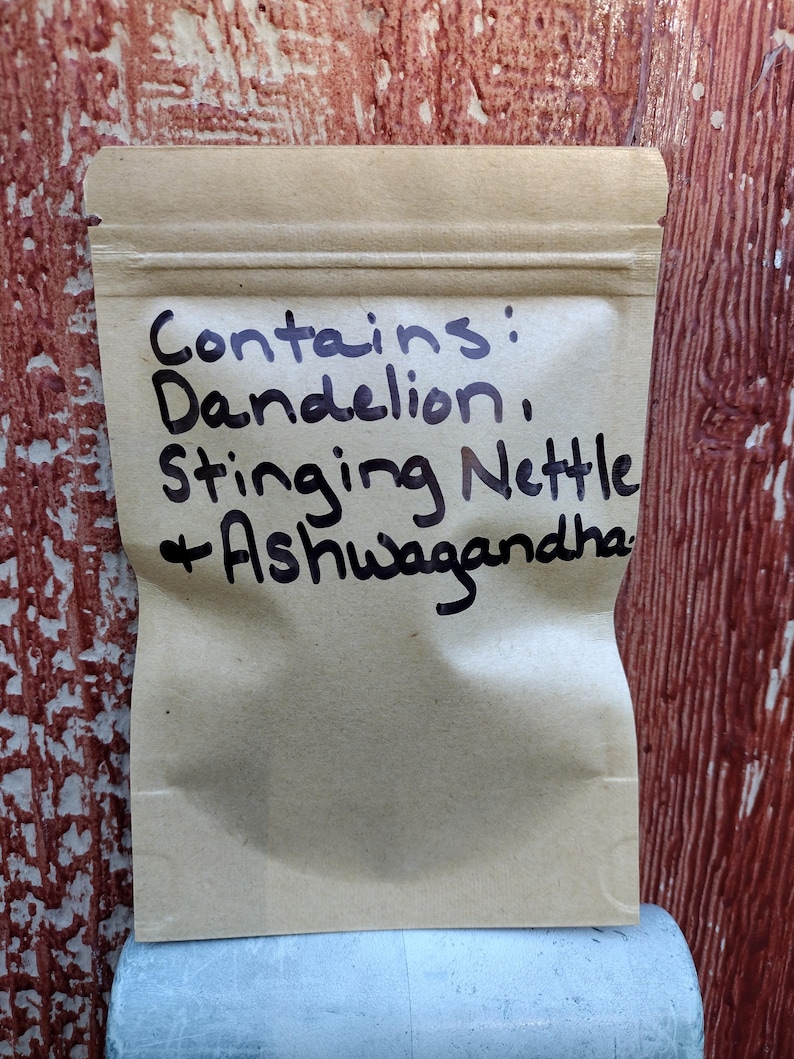 Pu&ograve; includere: Un sacchetto di carta marrone con un'etichetta scritta a mano che dice "Contiene: Tarassaco, Ortica + Ashwagandha."